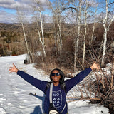 Woman wearing a navy blue No Wahala Crewneck sweatshirt with outstretched arms in a snowy elevation landscape with trees and mountains.