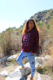 Woman wearing a maroon No Wahala Crewneck sweatshirt with hands in Ankara leggings pockets in mountain outdoors 